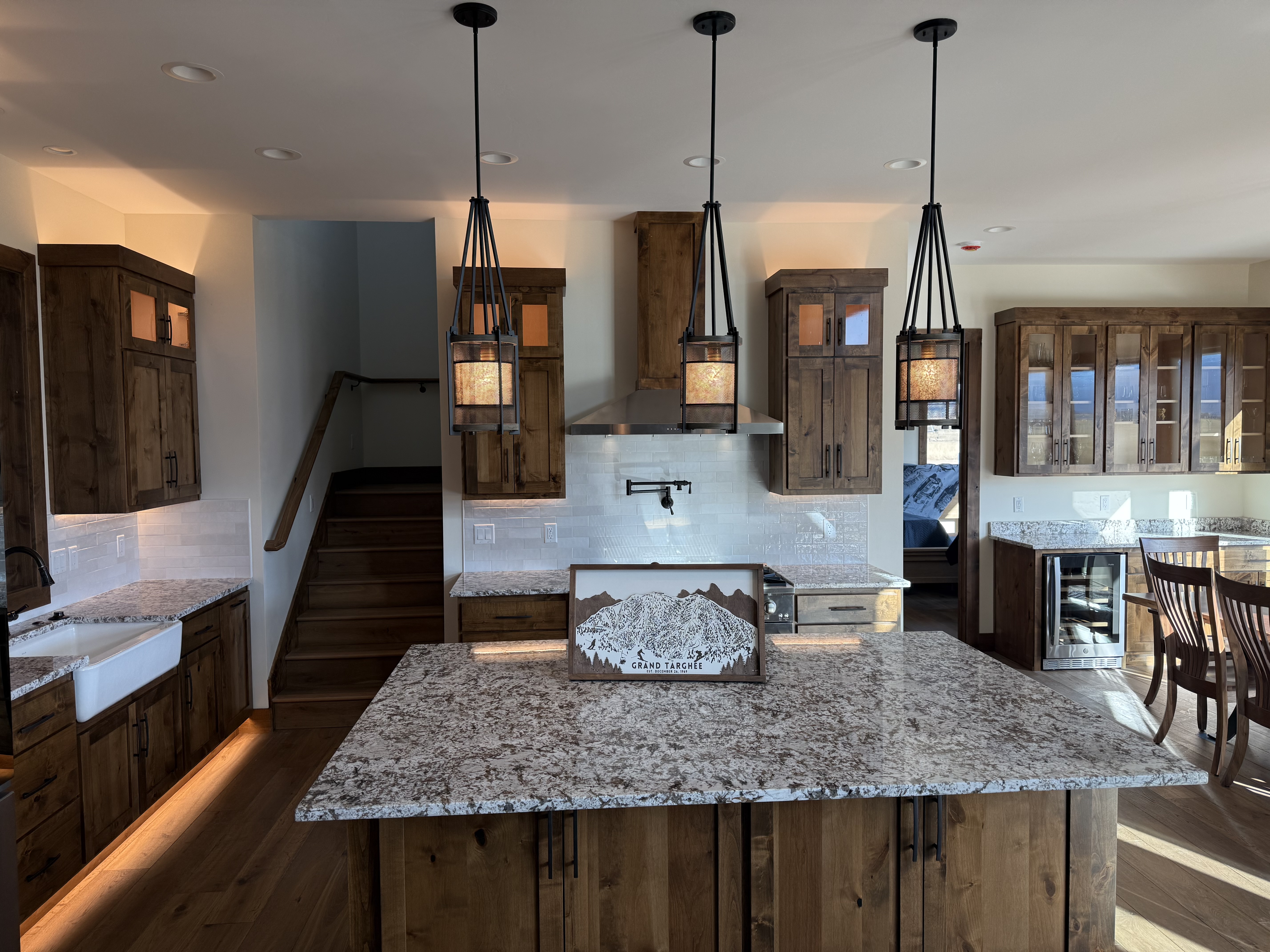 Modern kitchen featuring rustic wooden cabinets, granite countertops, stylish pendant lighting, and a scenic mountain view, reflecting luxury custom home design in Teton Valley.